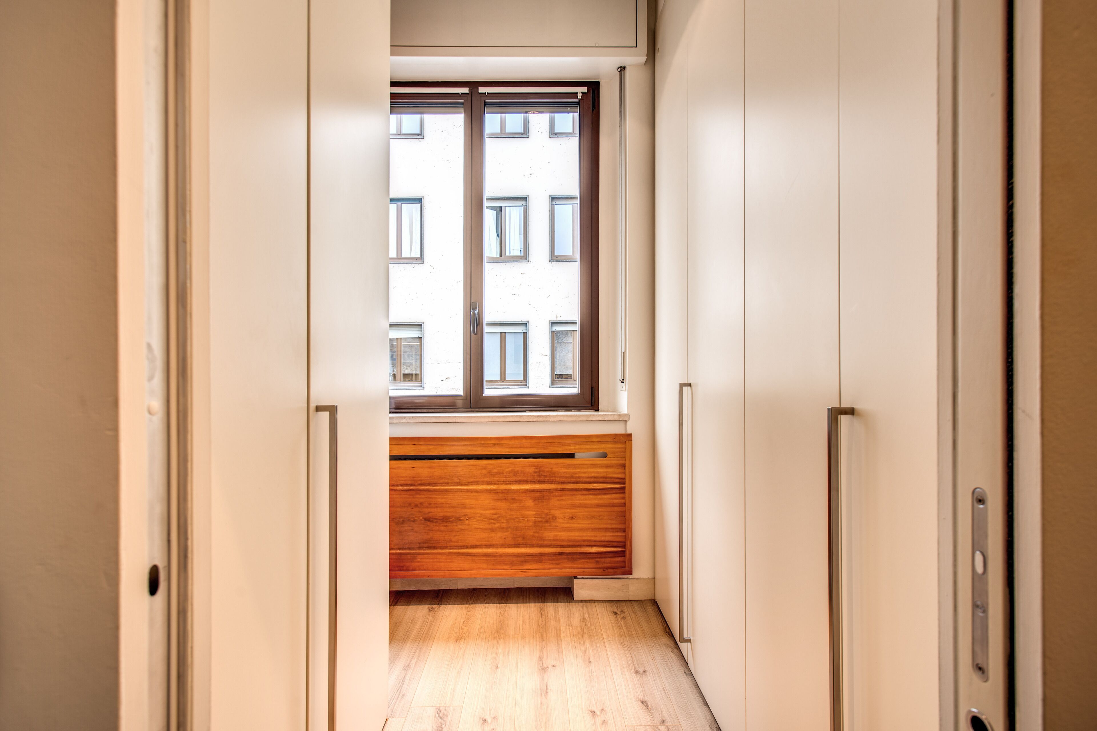 Un pasillo estrecho con un banco de madera, una ventana que da a un edificio y paredes blancas.