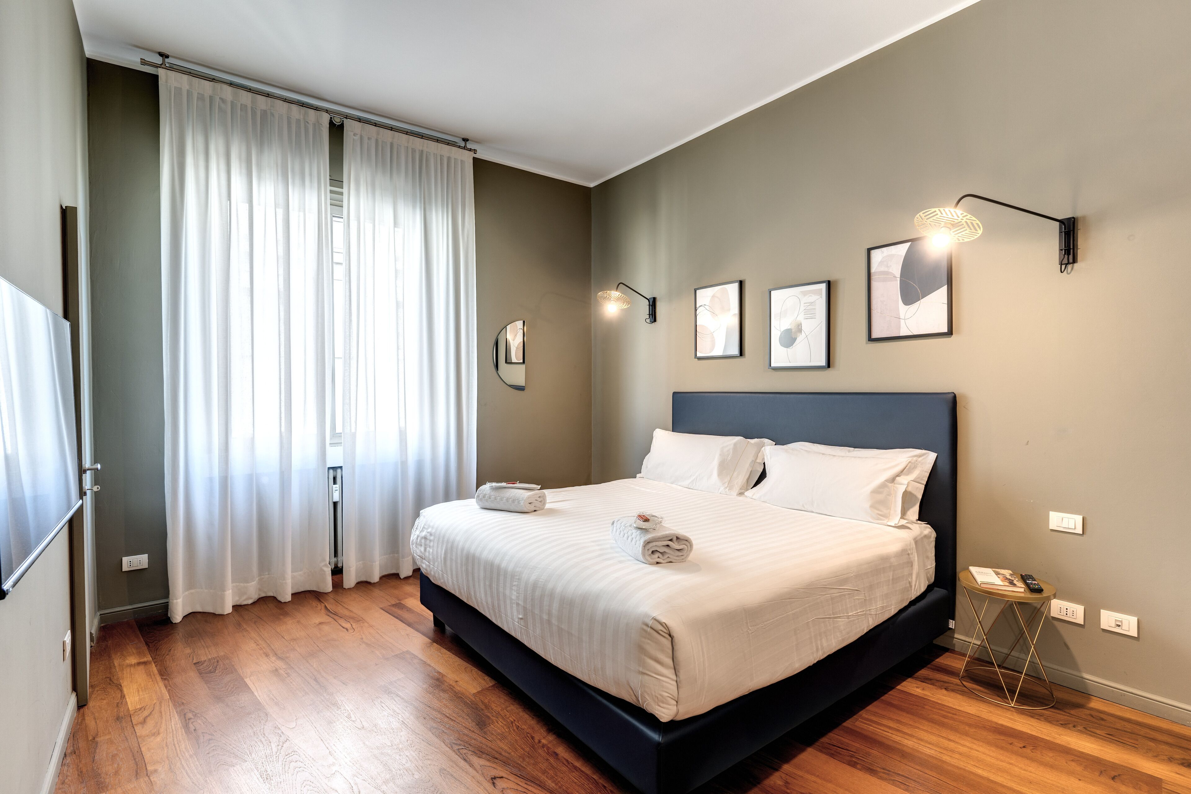 A modern bedroom with a large bed, a bedside table, a wall-mounted lamp, and a window with curtains.