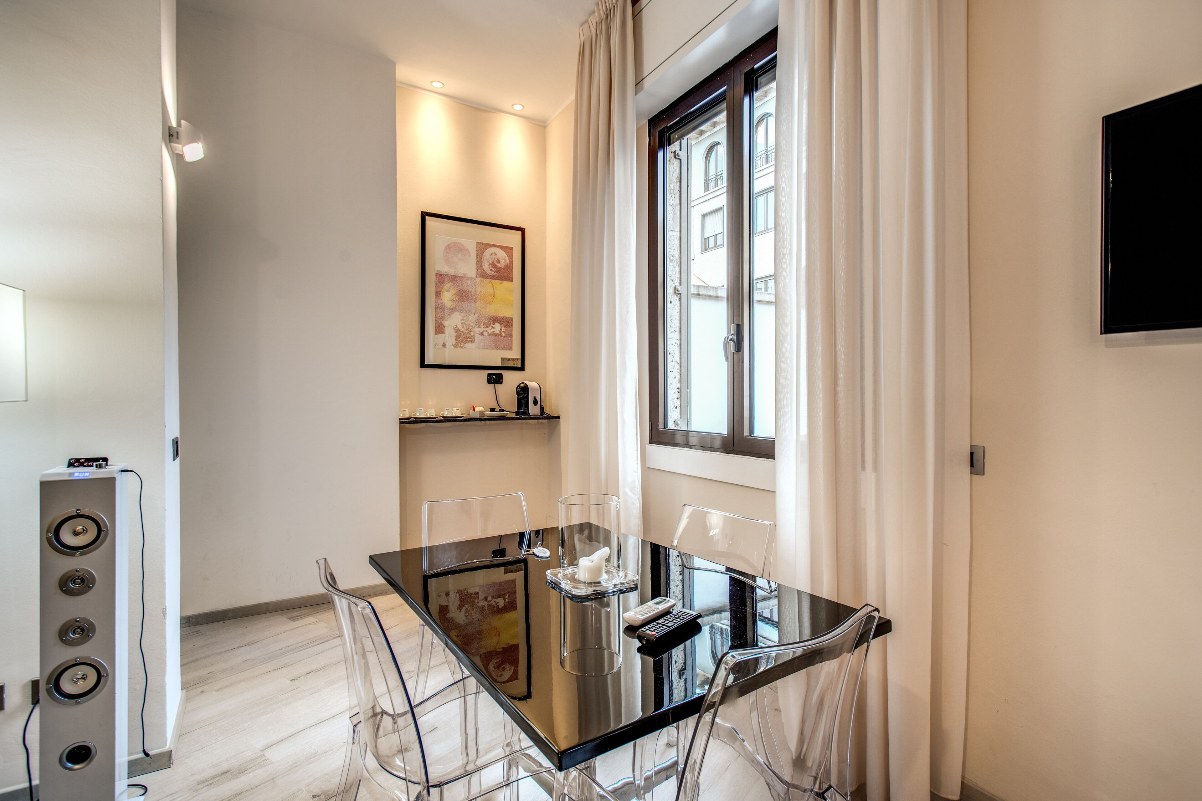 A modern dining area with a glass table, clear chairs, a mounted TV, and a window with curtains.