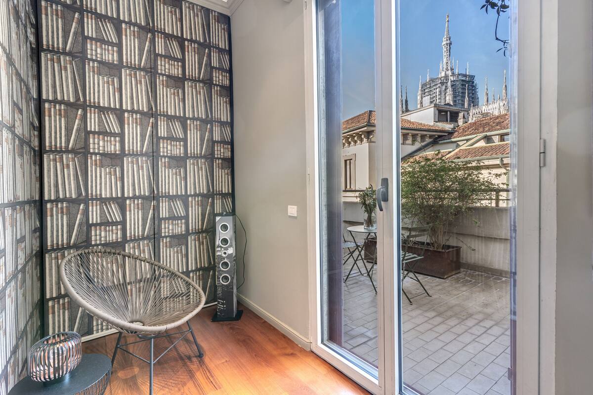 Une pièce comprenant une bibliothèque, un fauteuil, un haut-parleur et une porte-fenêtre coulissante donnant sur un patio extérieur avec vue sur un immeuble.