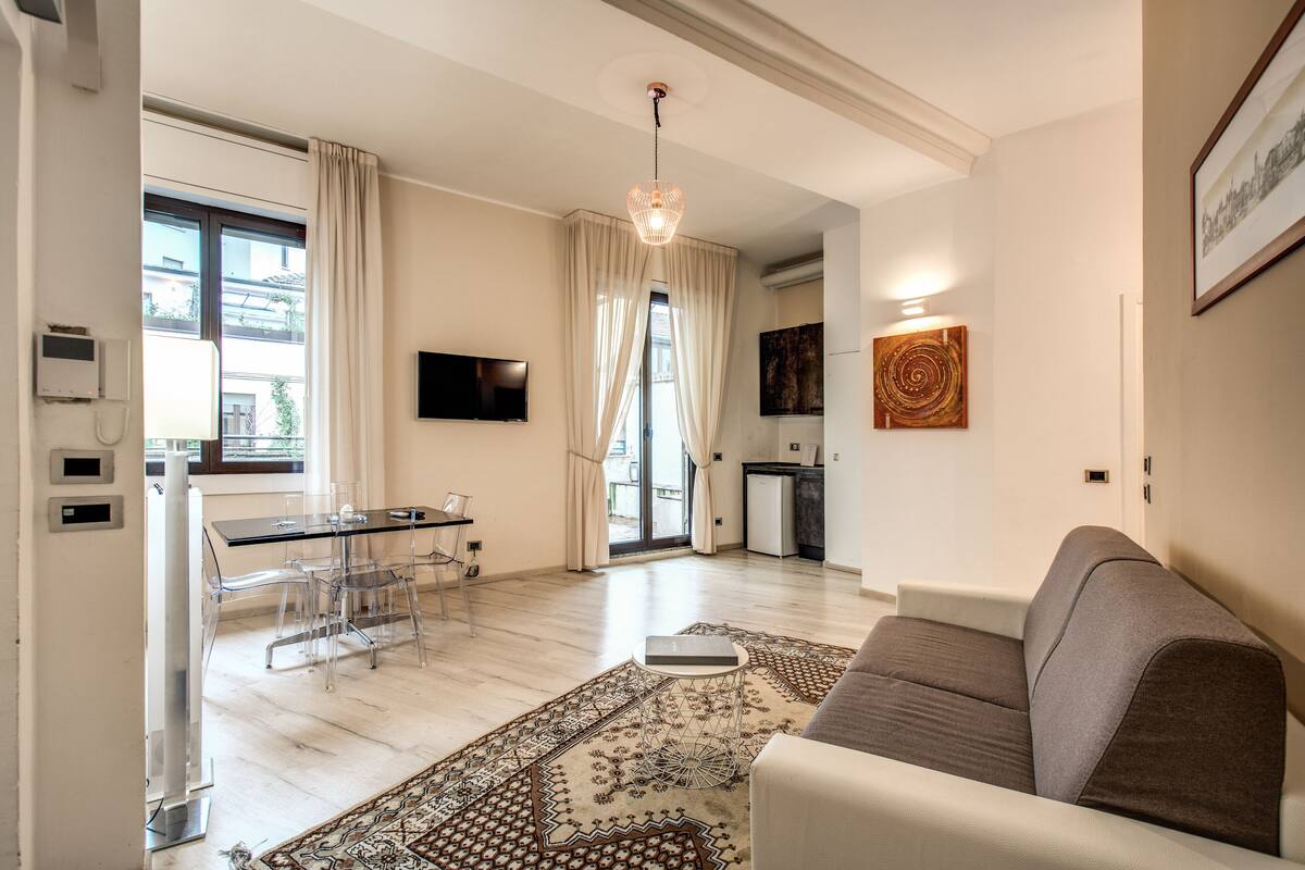 Un salon moderne avec un canapé, un coin repas avec une table et des chaises en verre, une cuisine à l’arrière-plan et une grande fenêtre avec des rideaux.