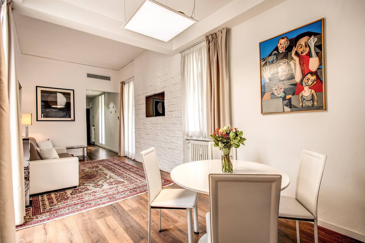 Un salon moderne avec un canapé blanc, une table de salle à manger ronde entourée de chaises, un tapis aux motifs rouges et beiges, et un tableau accroché au mur.