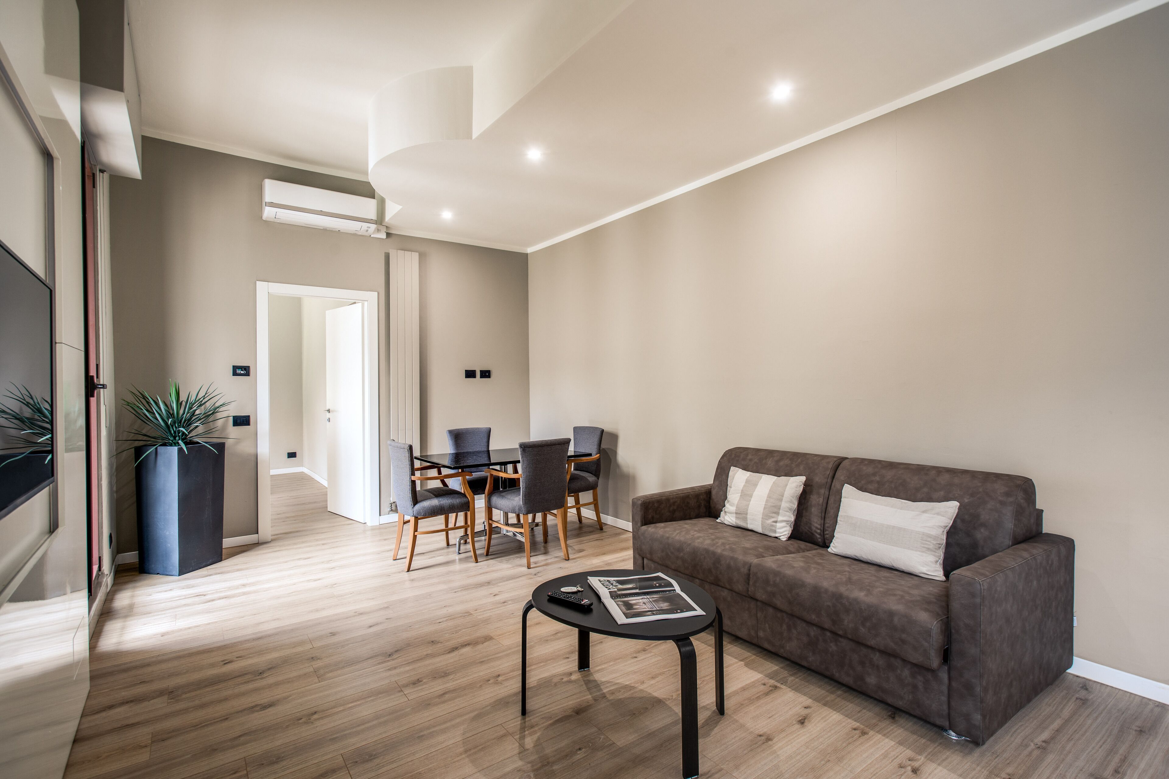 A modern living room with a brown sofa, a dining table with chairs, and a potted plant.