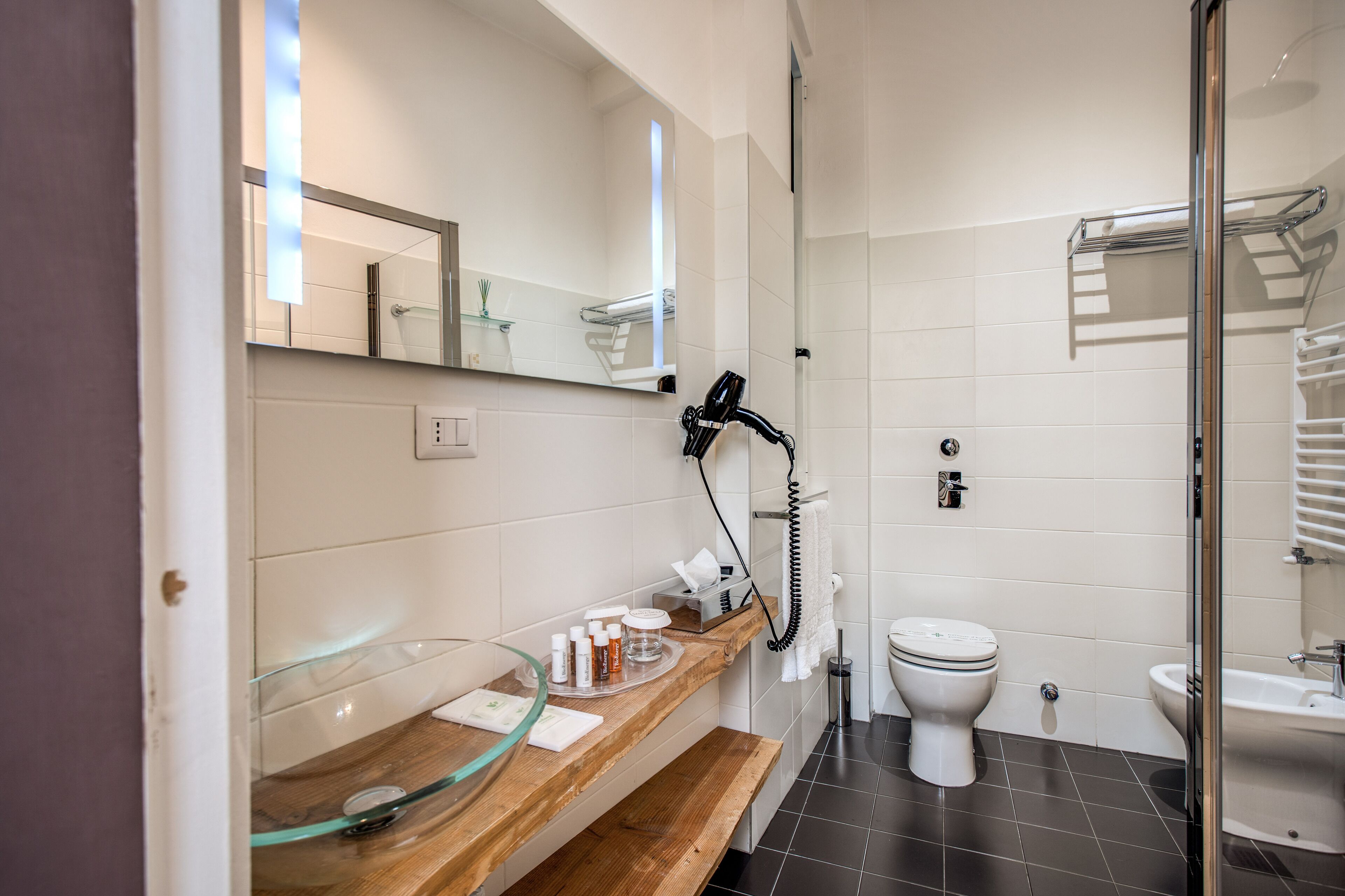 A modern bathroom with a glass shower enclosure, a wooden vanity with a glass bowl sink, a white toilet, and a white pedestal sink.