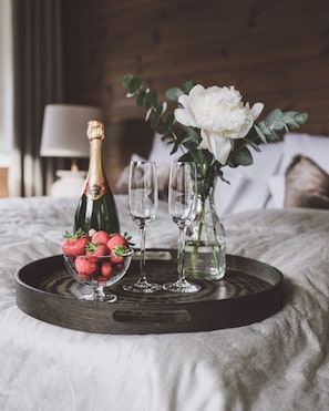 A tray with a bottle of champagne, a bowl of strawberries, and a vase with white flowers on a bed.