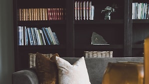 A living room with a dark wood bookshelf filled with books, a grey sofa with cushions, and a coffee table with a book and a vase. - Blåtind Boutique Hotel (Stordal)