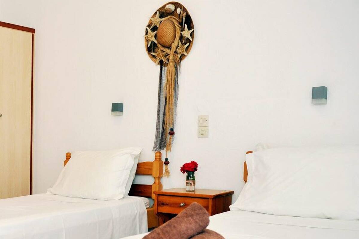 A hotel room with two beds, a wooden headboard, a wall-mounted decorative piece, a nightstand with a vase of red flowers, and a rolled towel on the bed.