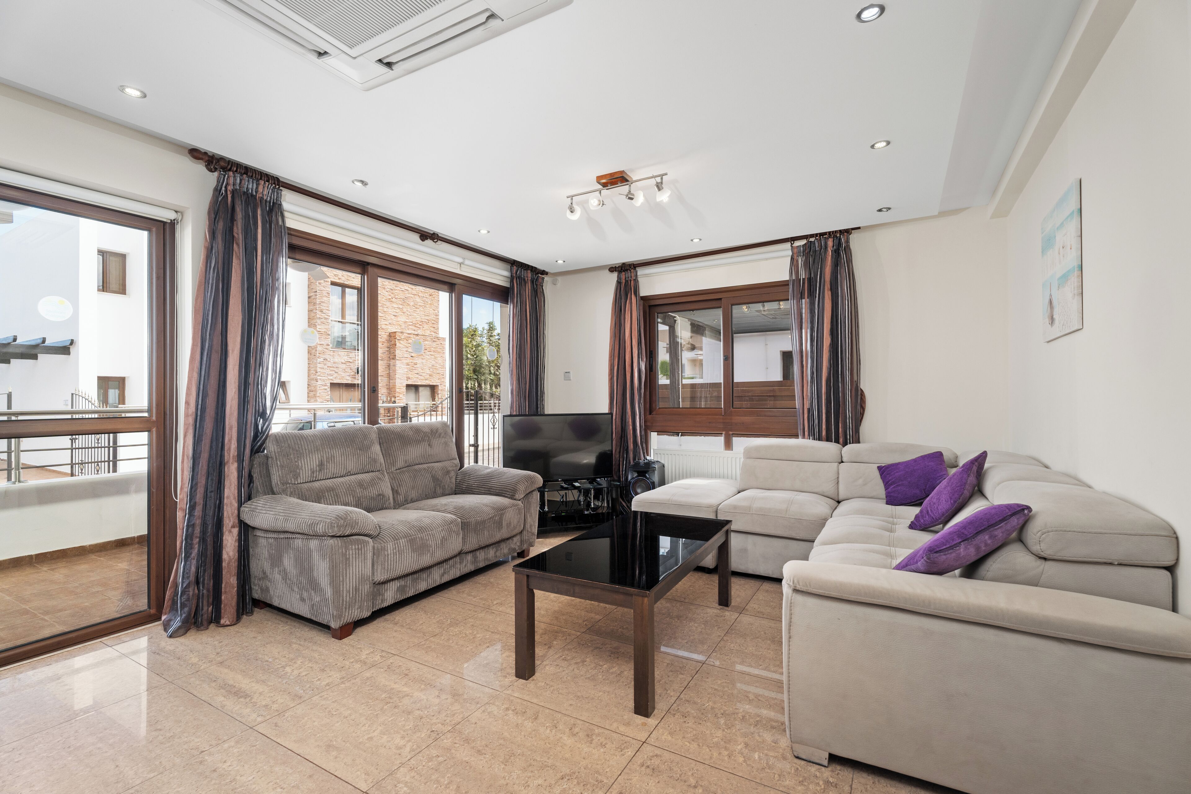 A modern living room with two sofas, a coffee table, and a TV. Large windows offer views of the outside.