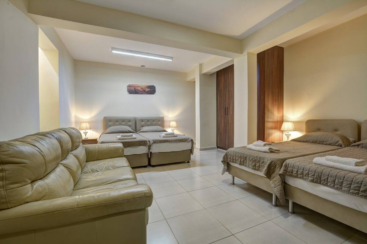 A hotel room with a sofa, two beds, a wooden wardrobe, and a painting on the wall.