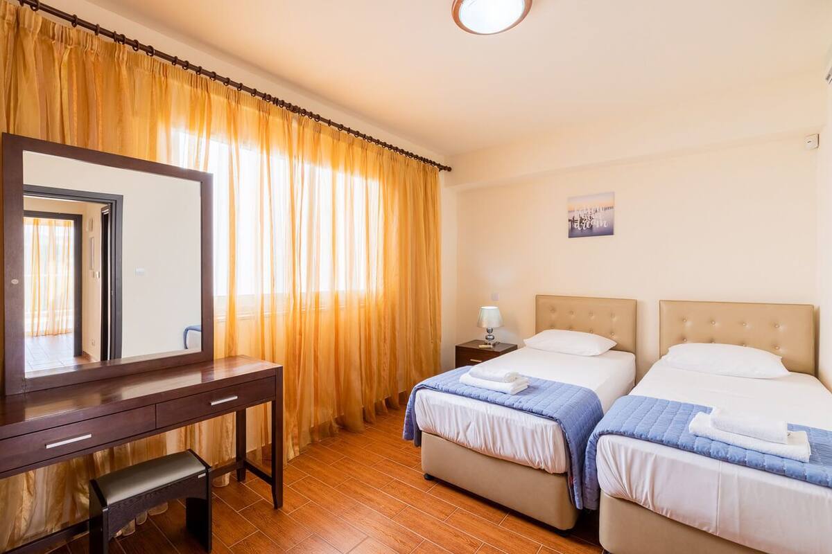 A hotel room with a wooden desk, two beds, a nightstand, and a large mirror.