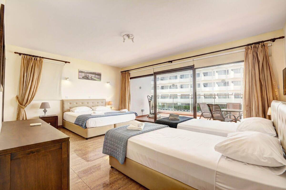 A hotel room with two beds, a desk, a chair, and a balcony with a view of the city.