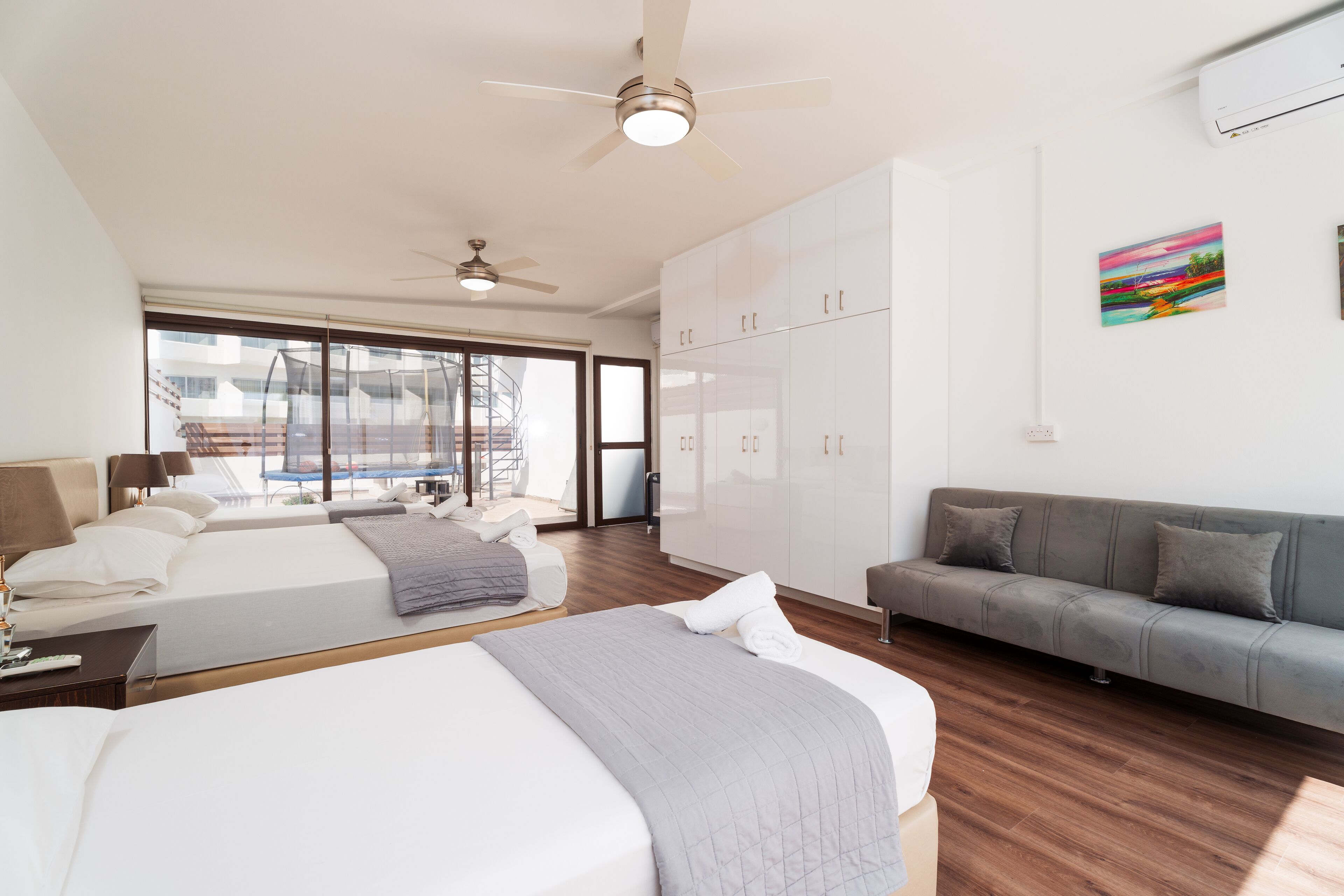 A hotel room with two beds, a gray sofa, a large window, and a ceiling fan.