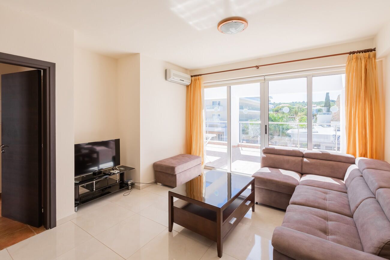 Ein modernes Wohnzimmer mit Flachbildfernseher, Glastisch, Sofa und einer Schiebetür zum Balkon.