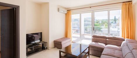 A modern living room with a flat-screen TV, a glass coffee table, a sofa, and a sliding glass door leading to a balcony.
