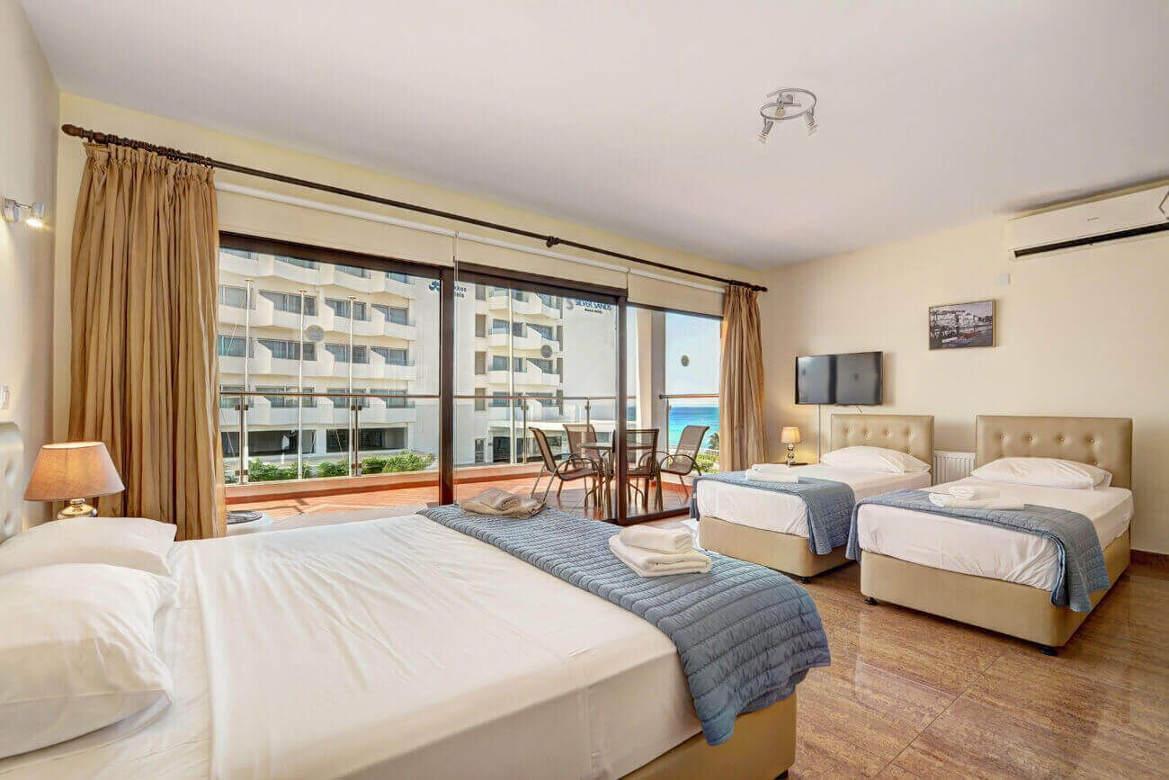 A hotel room with two beds, a balcony with a table and chairs, and a large window offering a view of the city.