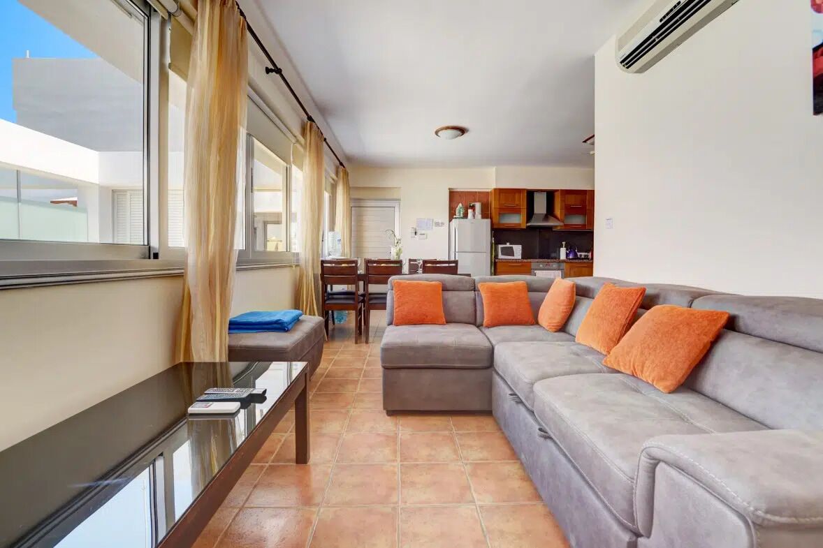 Un salón moderno con un sofá gris, cojines naranja, una mesa de centro de vidrio y vistas a la ciudad a través de amplias ventanas.