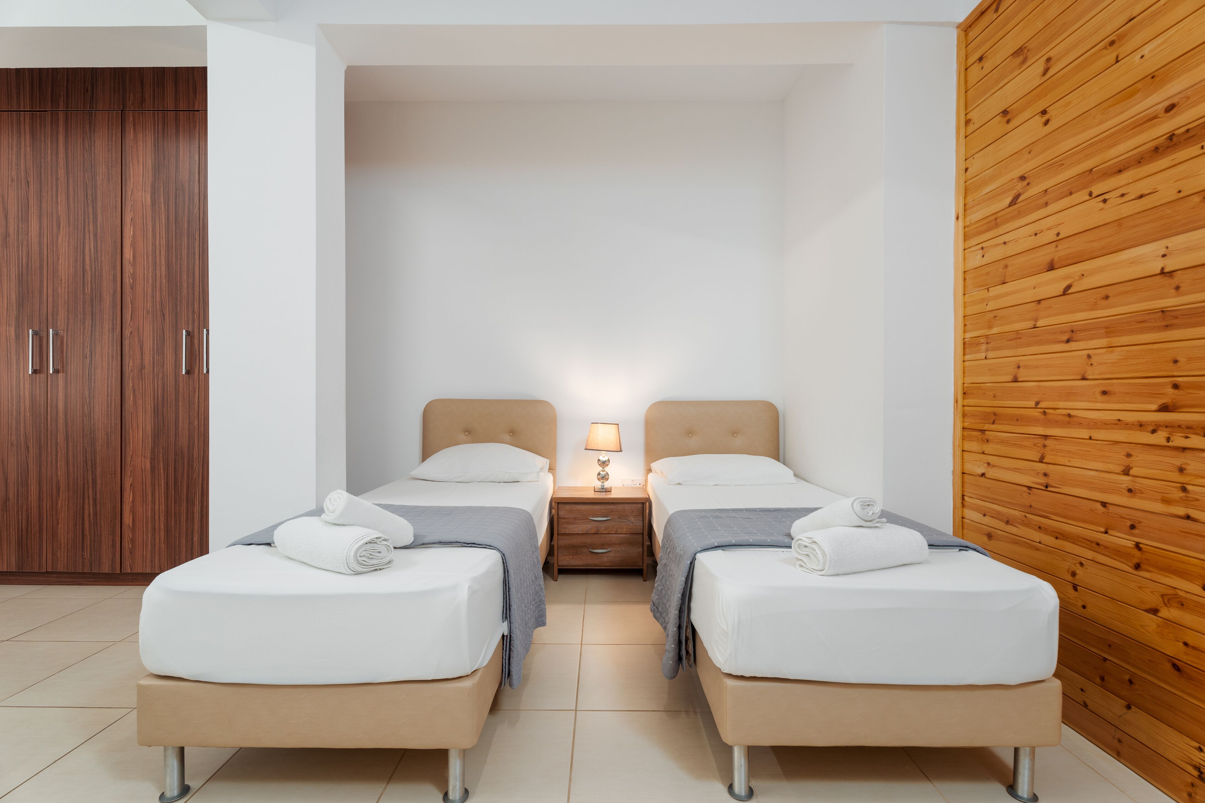 A modern hotel room with two single beds, a wooden wardrobe, a nightstand with a lamp, and a wooden panel wall.