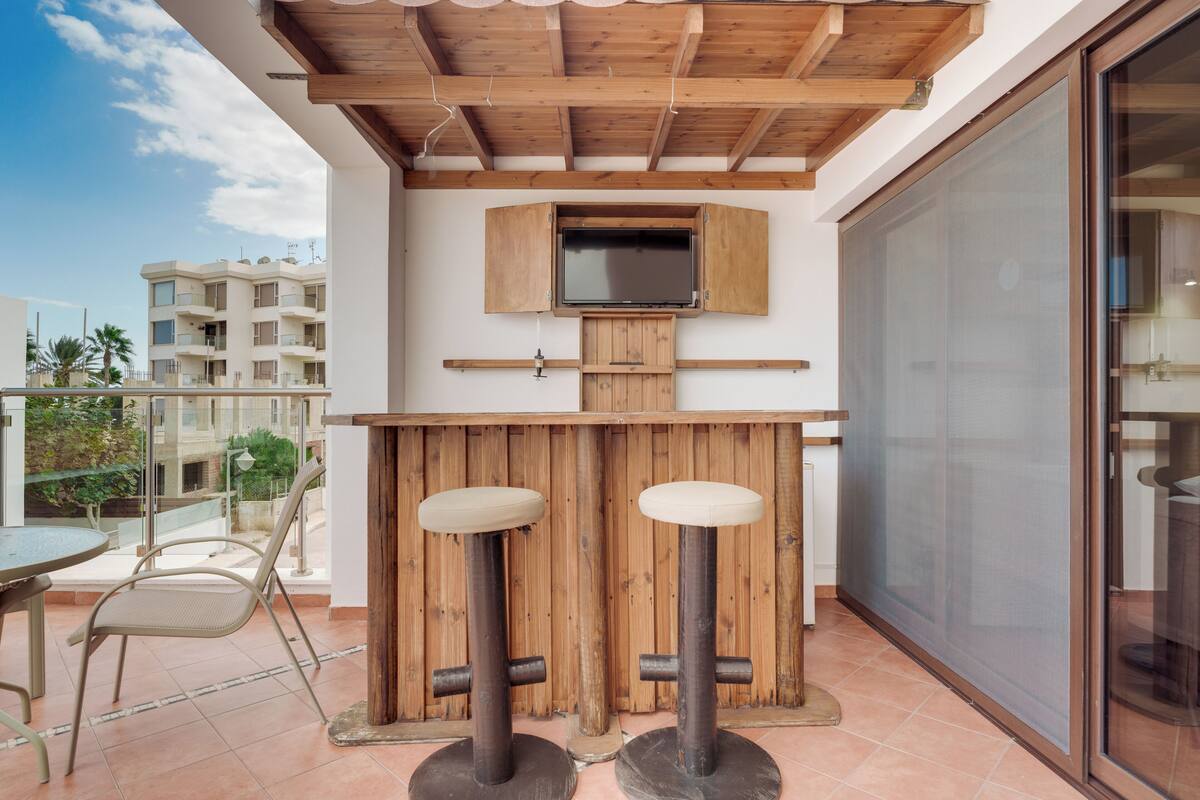 A balcony with a wooden bar, two stools, a television mounted on a wooden cabinet, and a glass door.