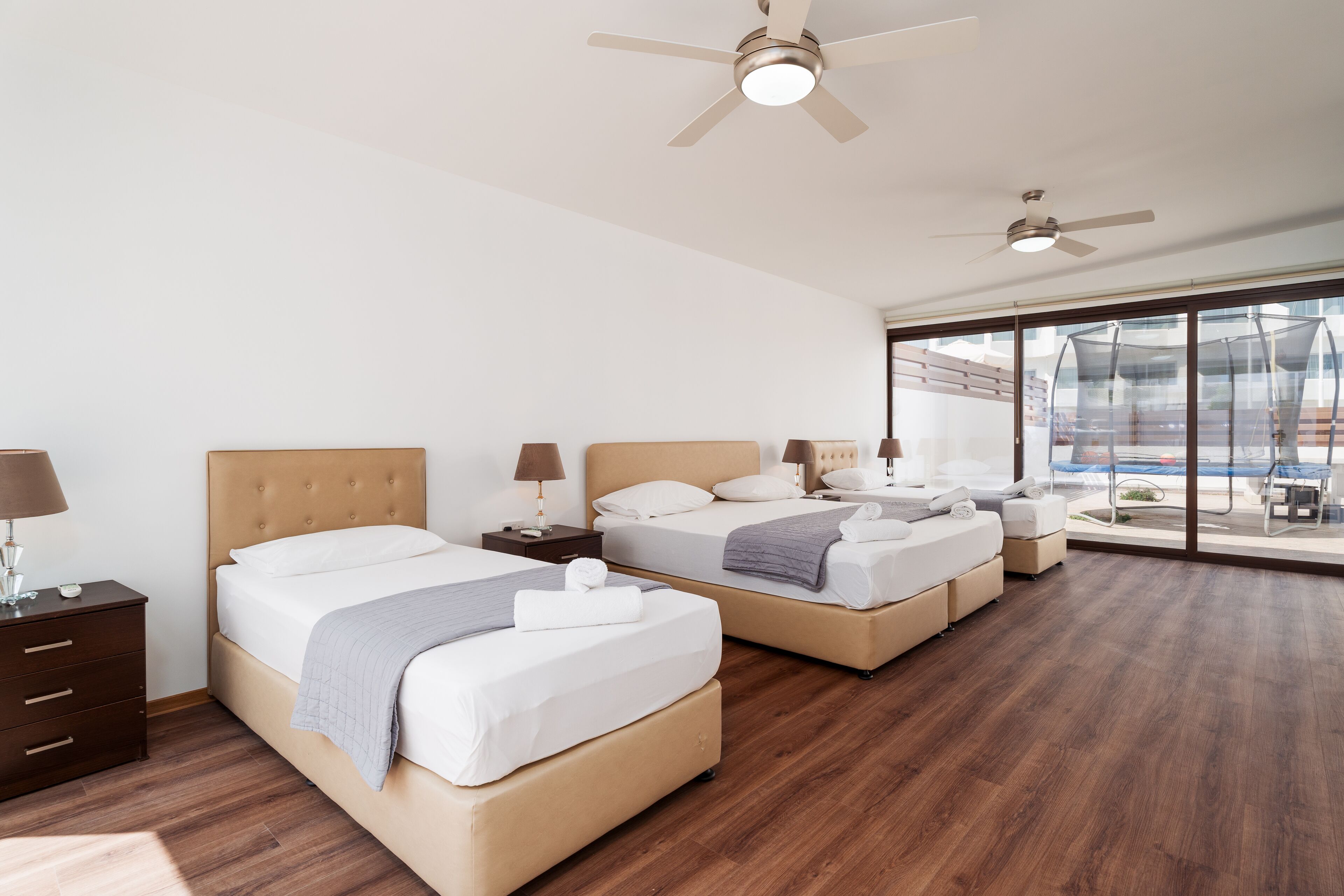 A modern hotel room with two beds, wooden flooring, ceiling fans, and large windows offering a view of the outdoors.