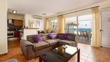 Un salón con sofá, una mesa de centro y un comedor con sillas y una mesa. Hay un ventanal corredero que da a un balcón con vistas al mar.