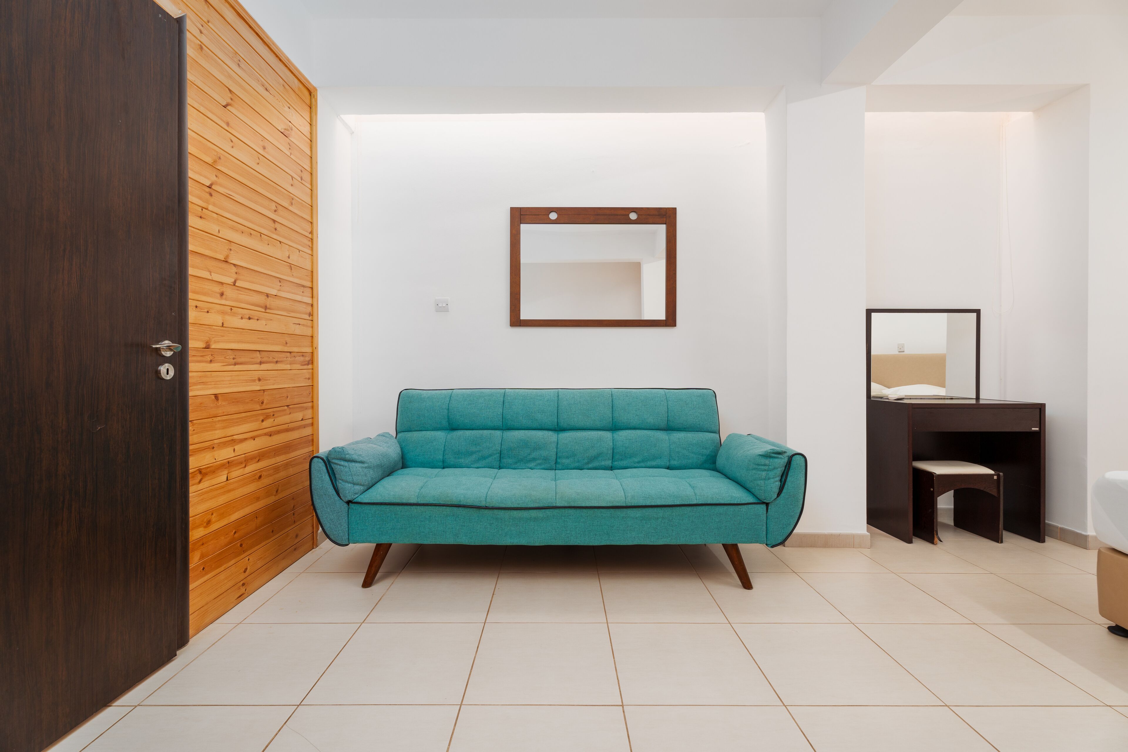 A room with a teal sofa, a wooden wall, a small desk with a chair, and a mirror on the wall.
