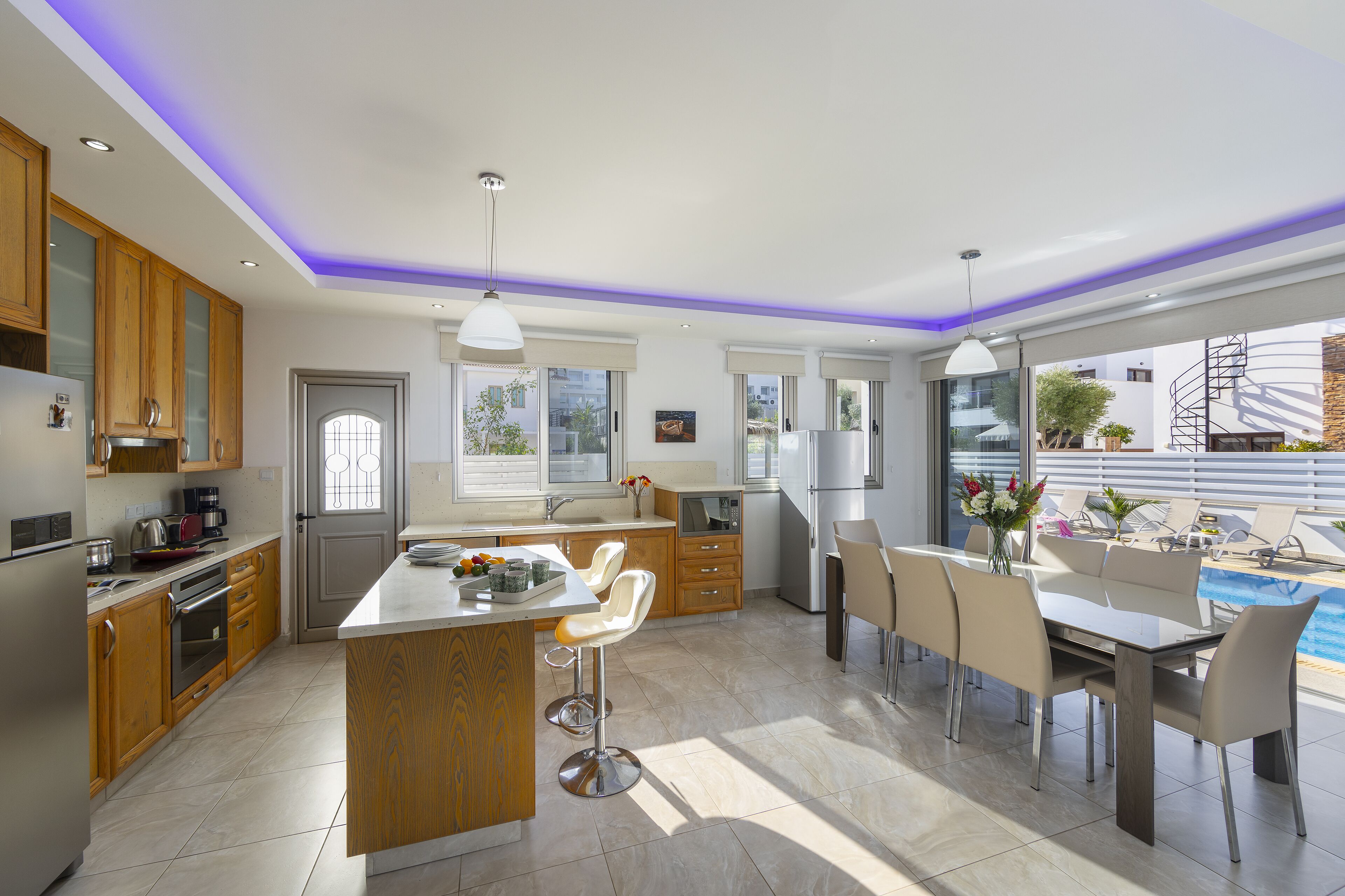 A modern kitchen with wooden cabinets, a central island, bar stools, and a dining area with a pool view.