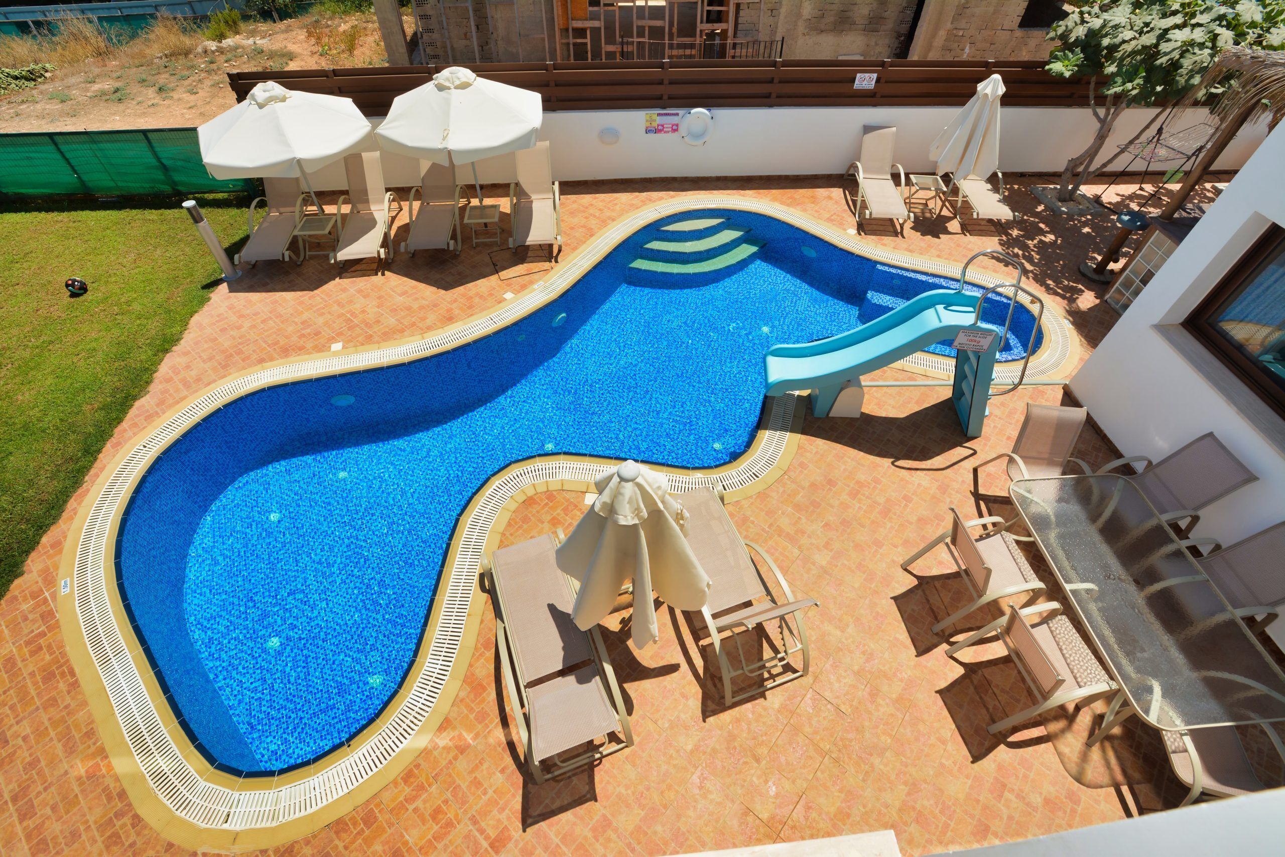 Une piscine avec un toboggan, entourée de chaises longues et de parasols, et une table avec des chaises.