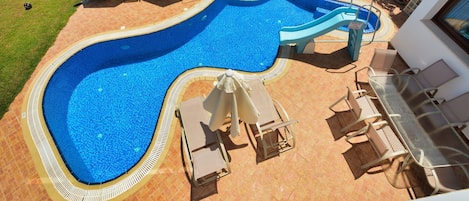 A swimming pool with a slide, surrounded by lounge chairs and umbrellas, and a table with chairs.