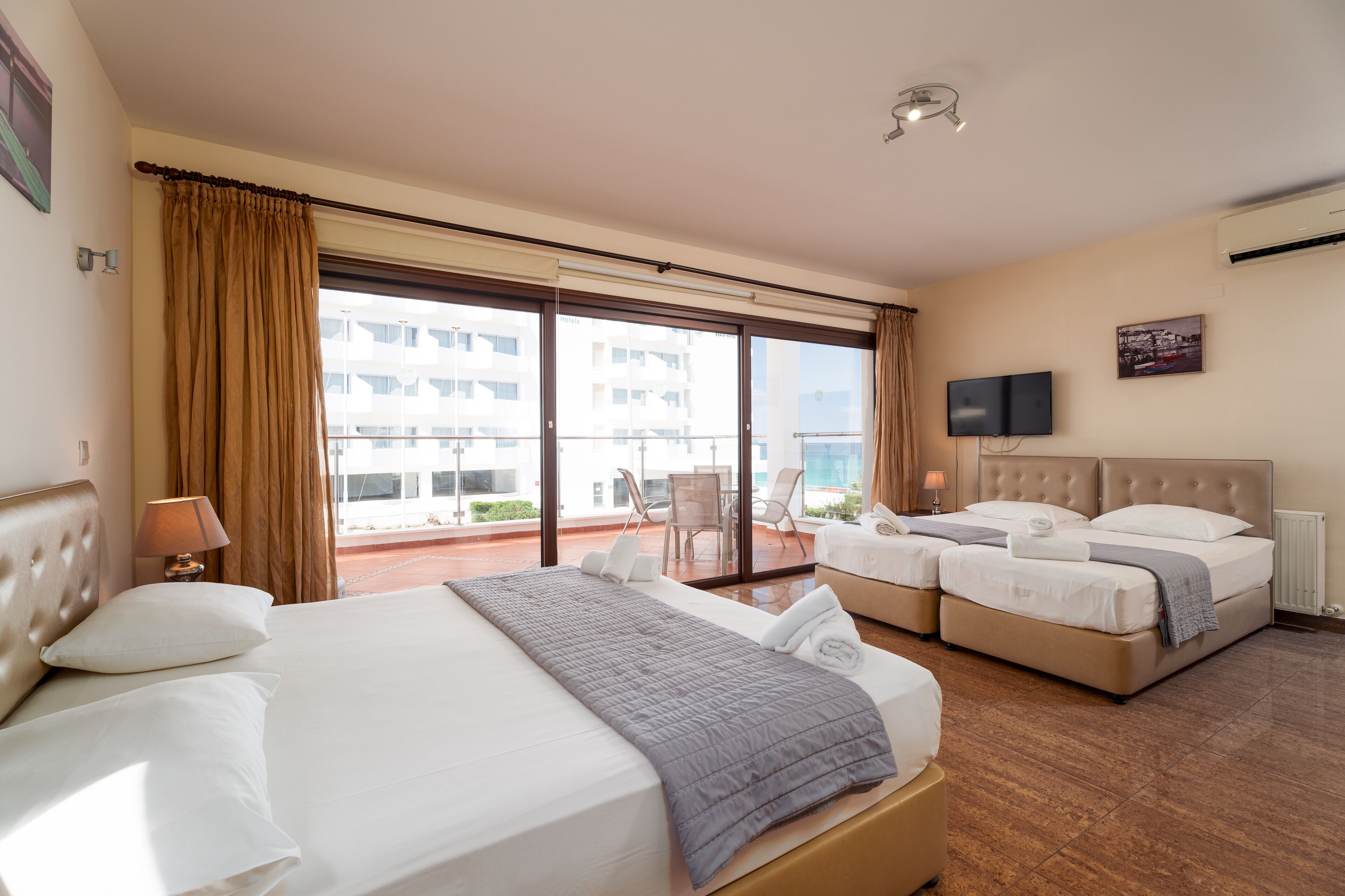 Une chambre d’hôtel avec un grand lit, deux petits lits, un balcon avec des chaises et un téléviseur fixé au mur.