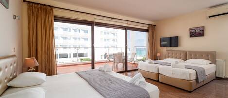 Une chambre d’hôtel avec un grand lit, deux petits lits, un balcon avec des chaises et un téléviseur fixé au mur.
