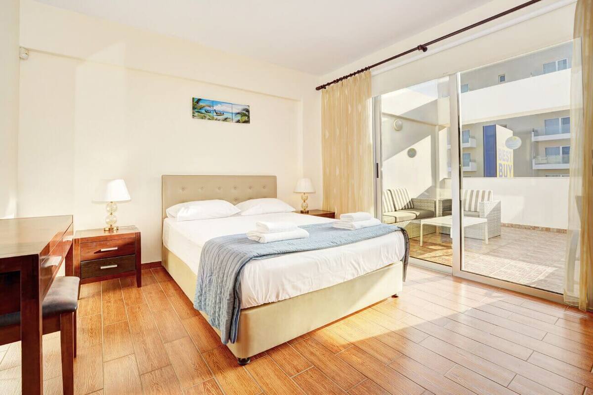 A hotel room with a bed, bedside tables, a desk, and a balcony with outdoor seating.