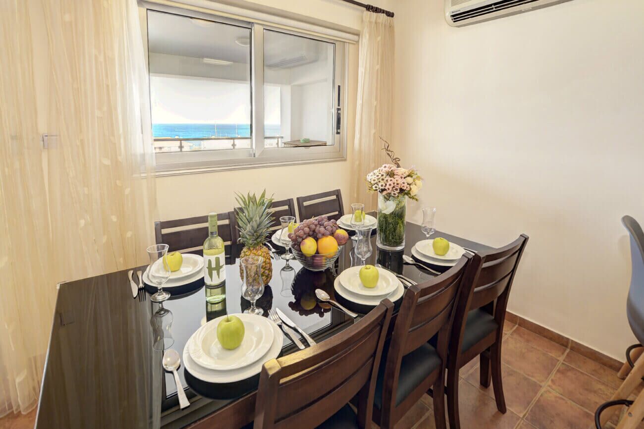 Un comedor con una mesa dispuesta para cuatro, una ventana con vista al mar y un jarrón con flores sobre la mesa.