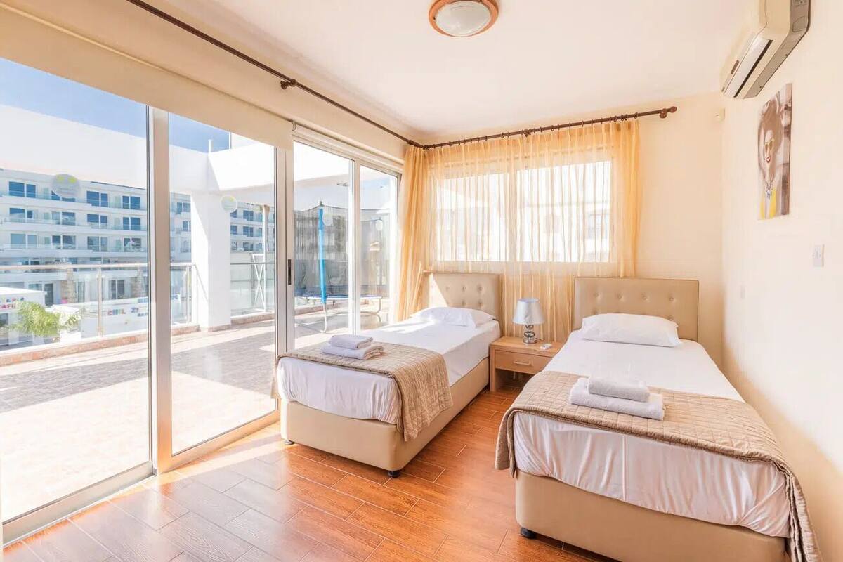 A hotel room with two beds, a large window with a view of buildings, and a ceiling fan.