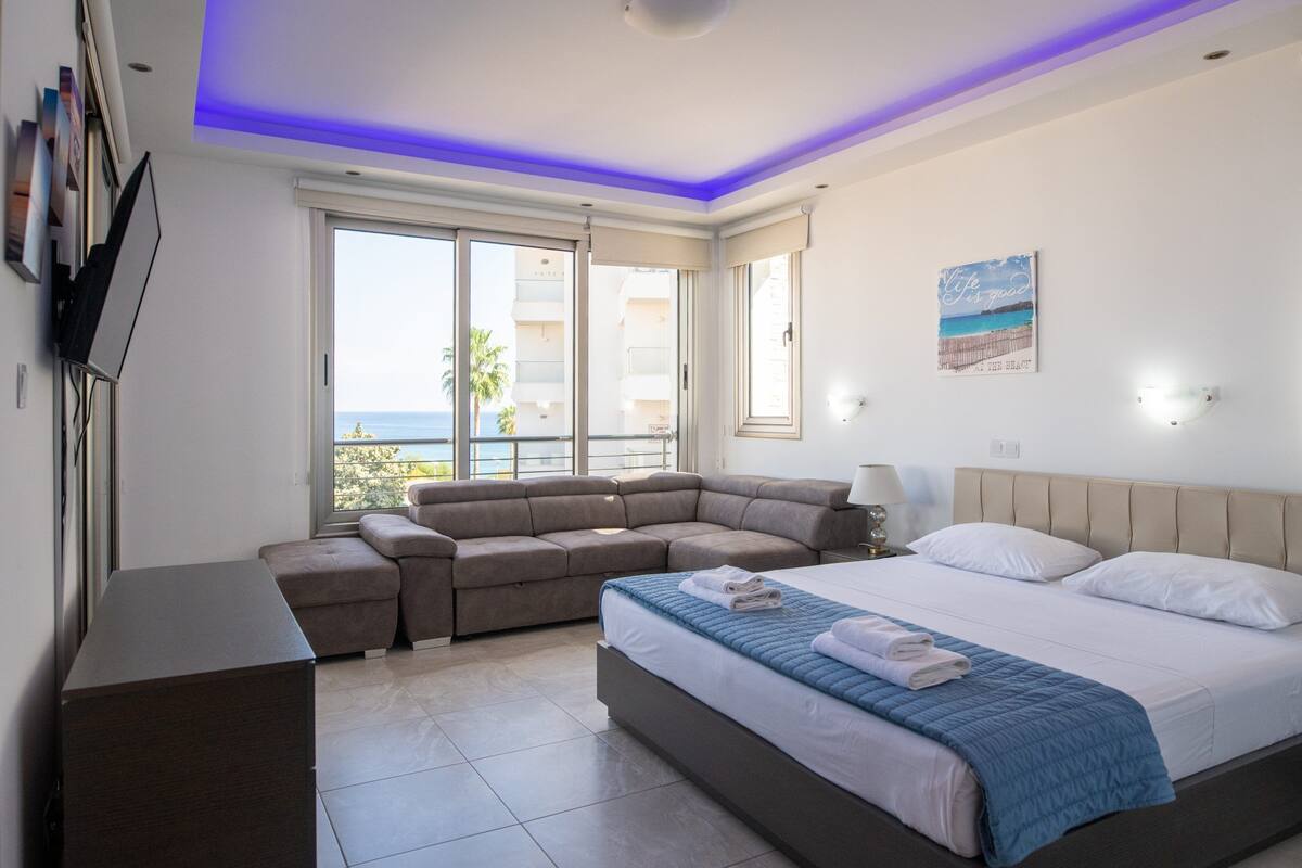A modern bedroom with a large bed, a sofa, a TV, and a view of the sea.