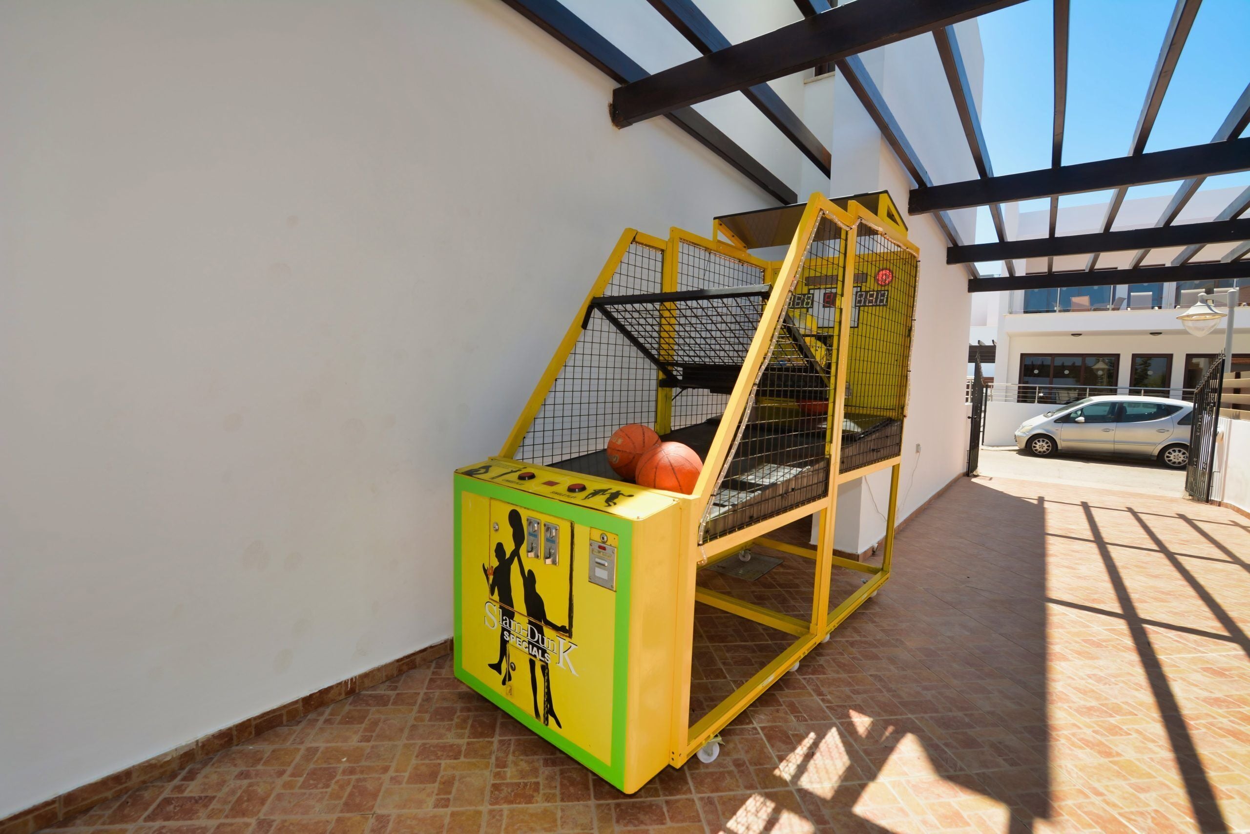 En basketballkurv med en gul og grøn indhegning, en basketball, og en bil parkeret i baggrunden.