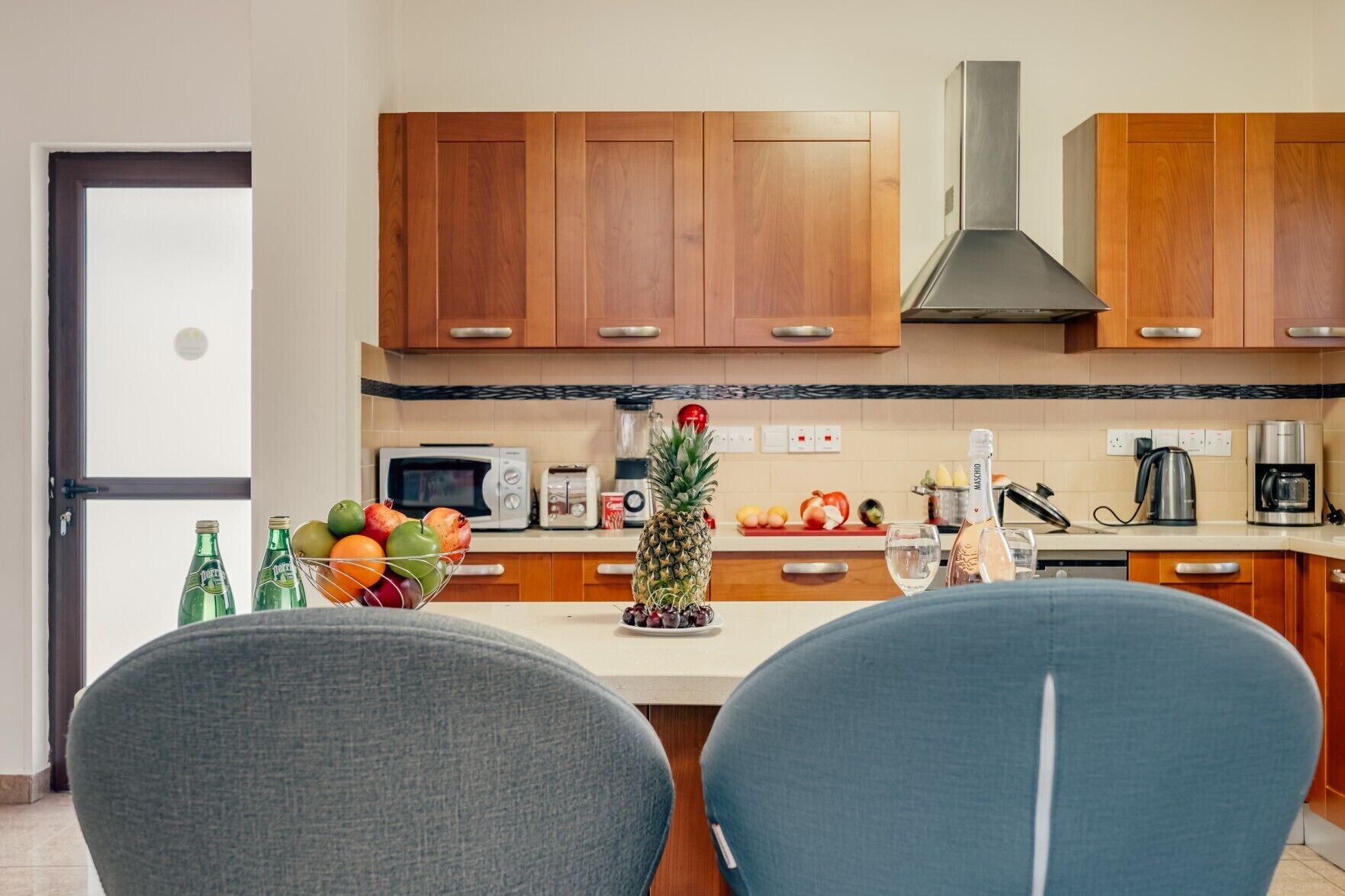 Una cucina moderna con mobili in legno, un forno a microonde, un tostapane e una macchina da caffè. Sul bancone c'è una ciotola di frutta e una bottiglia di vino.