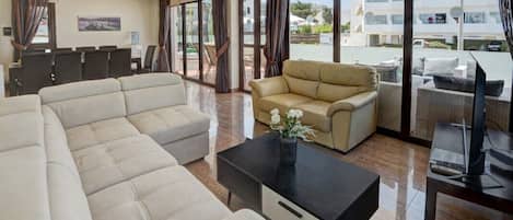 Una habitación de hotel moderna con un sofá grande, otro más pequeño, una mesita de centro y un escritorio con una computadora. La habitación tiene ventanas amplias con cortinas, vistas al exterior y un balcón con mobiliario exterior.