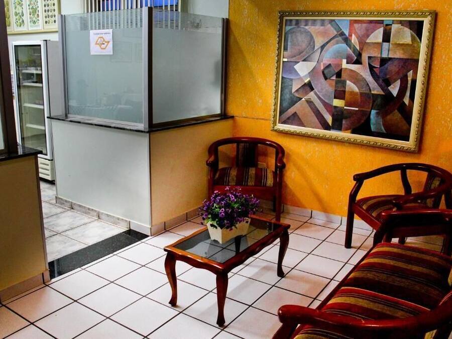 A room with a tiled floor, a glass-top coffee table with a flower arrangement, two wooden chairs, a sofa, a refrigerator, and a framed abstract painting on the wall.