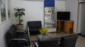 A reception area with a reception desk, black leather chairs, a potted plant, a glass coffee table with yellow flowers, a television on a wooden stand, and a glass display cabinet with various items. - Alpha Ville Hotel (Assis)
