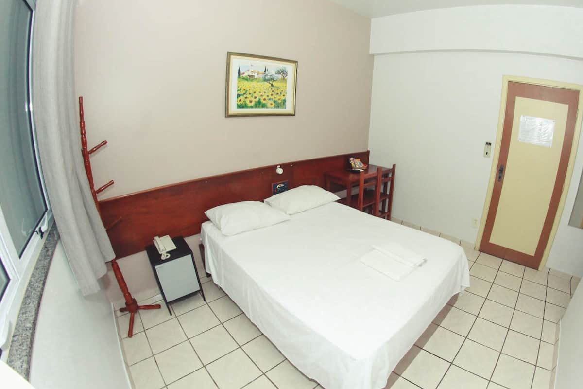 Habitación de hotel con cama, mesita de noche, silla, puerta y un cuadro en la pared.