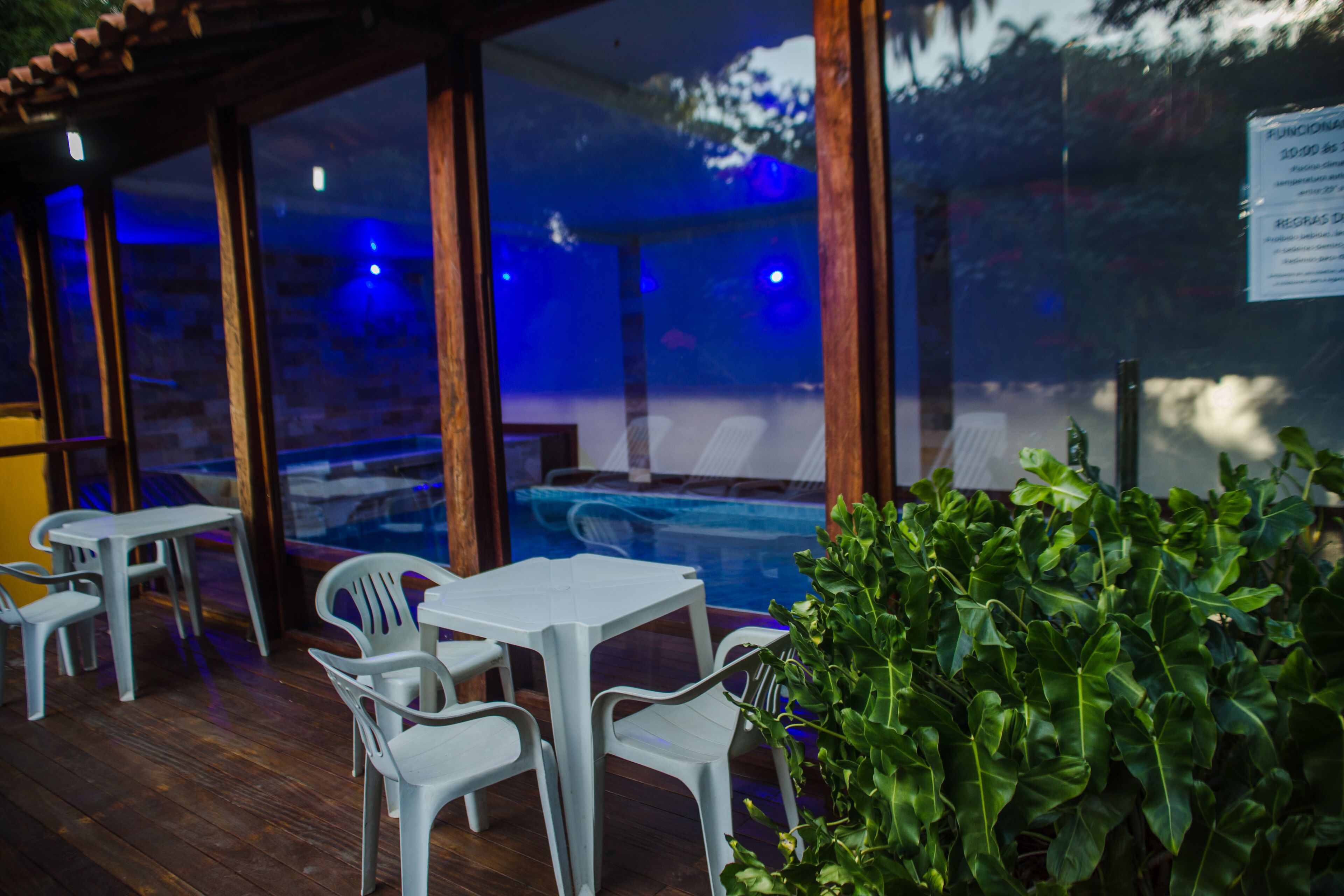 An outdoor seating area with plastic chairs and tables, a glass enclosure, and a pool in the background.