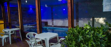 An outdoor seating area with plastic chairs and tables, a glass enclosure, and a pool in the background.