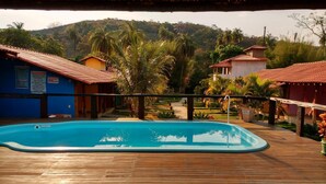 Une terrasse en bois avec une piscine, entourée de palmiers et d’immeubles aux toits recouverts de tuiles rouges.