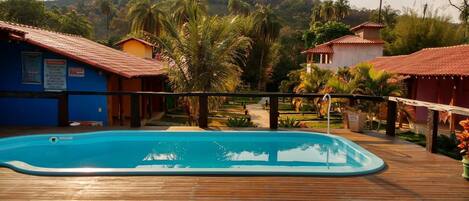 A wooden deck with a swimming pool, surrounded by palm trees and buildings with red-tiled roofs.