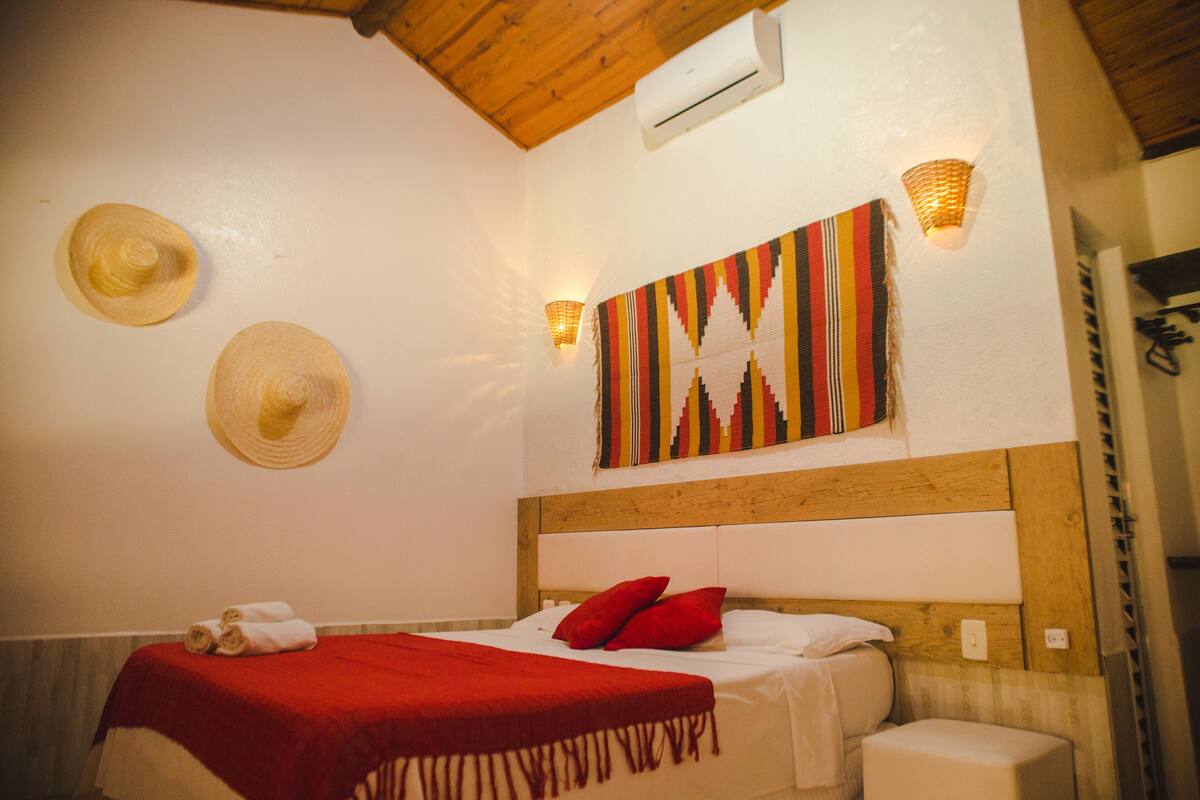 A bedroom with a wooden headboard, a red bedspread, two red pillows, and two straw hats hanging on the wall.