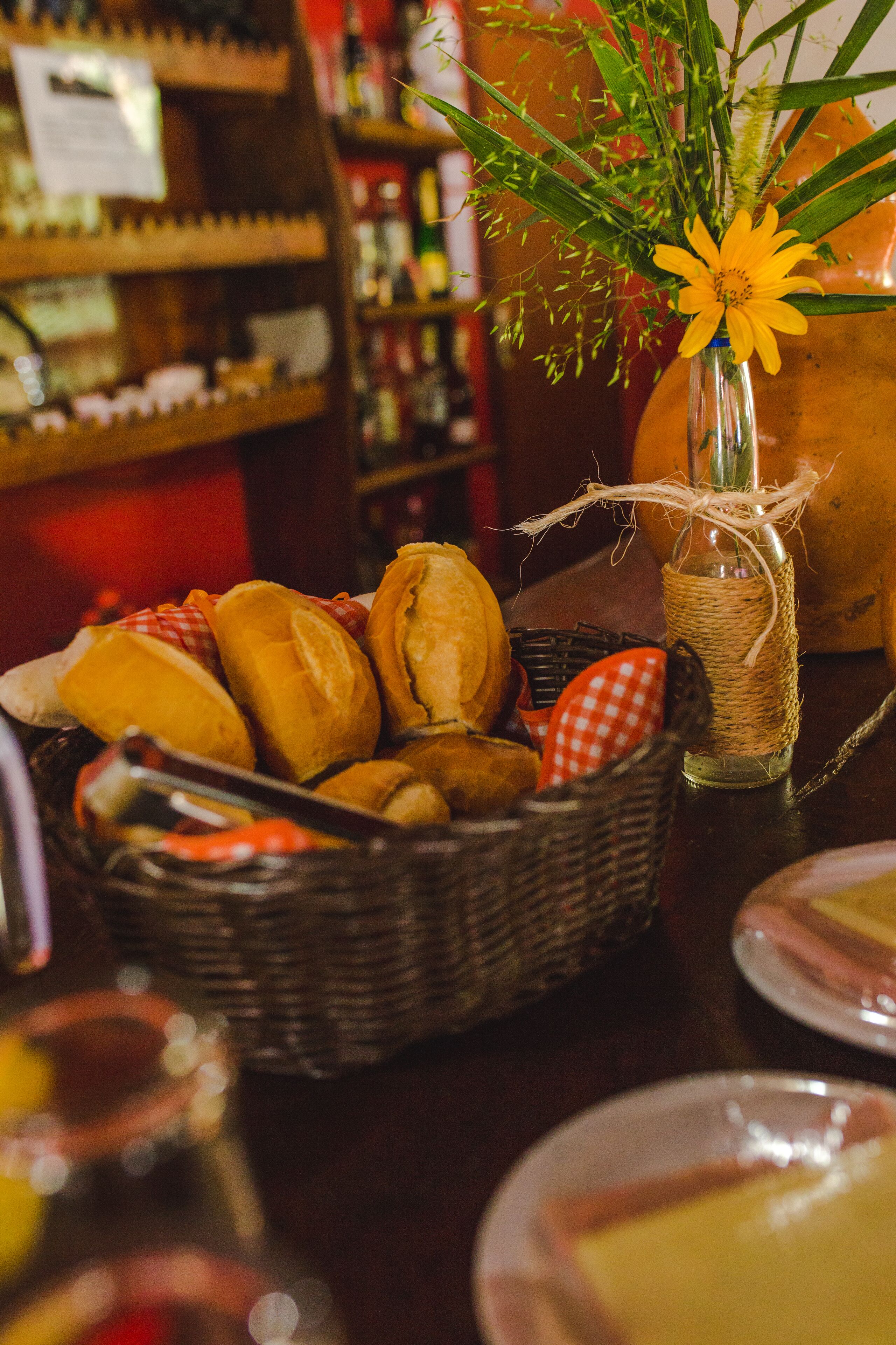 A table with a basket of bread rolls, a small vase with a yellow flower, and a wicker basket with a red and white checkered cloth.
