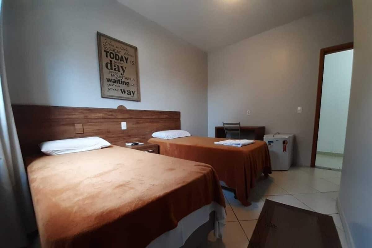 Ett hotellrum med två sängar, en sänggavel i trä, ett litet bord, en stol och ett kylskåp.