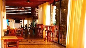 Una habitación amplia con suelo de madera, una zona de comedor con mesas y sillas, y ventanas grandes con cortinas.
