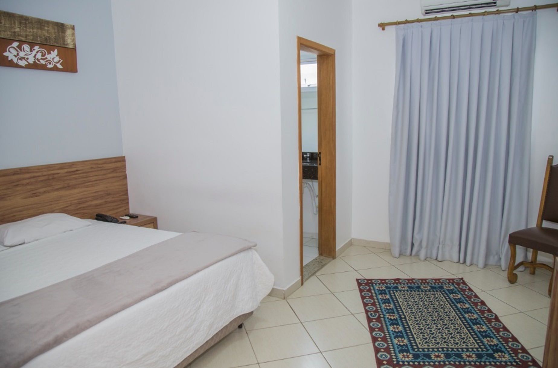 A bedroom with a bed, a nightstand, a wooden headboard, a bathroom door, a window with curtains, and a patterned rug.