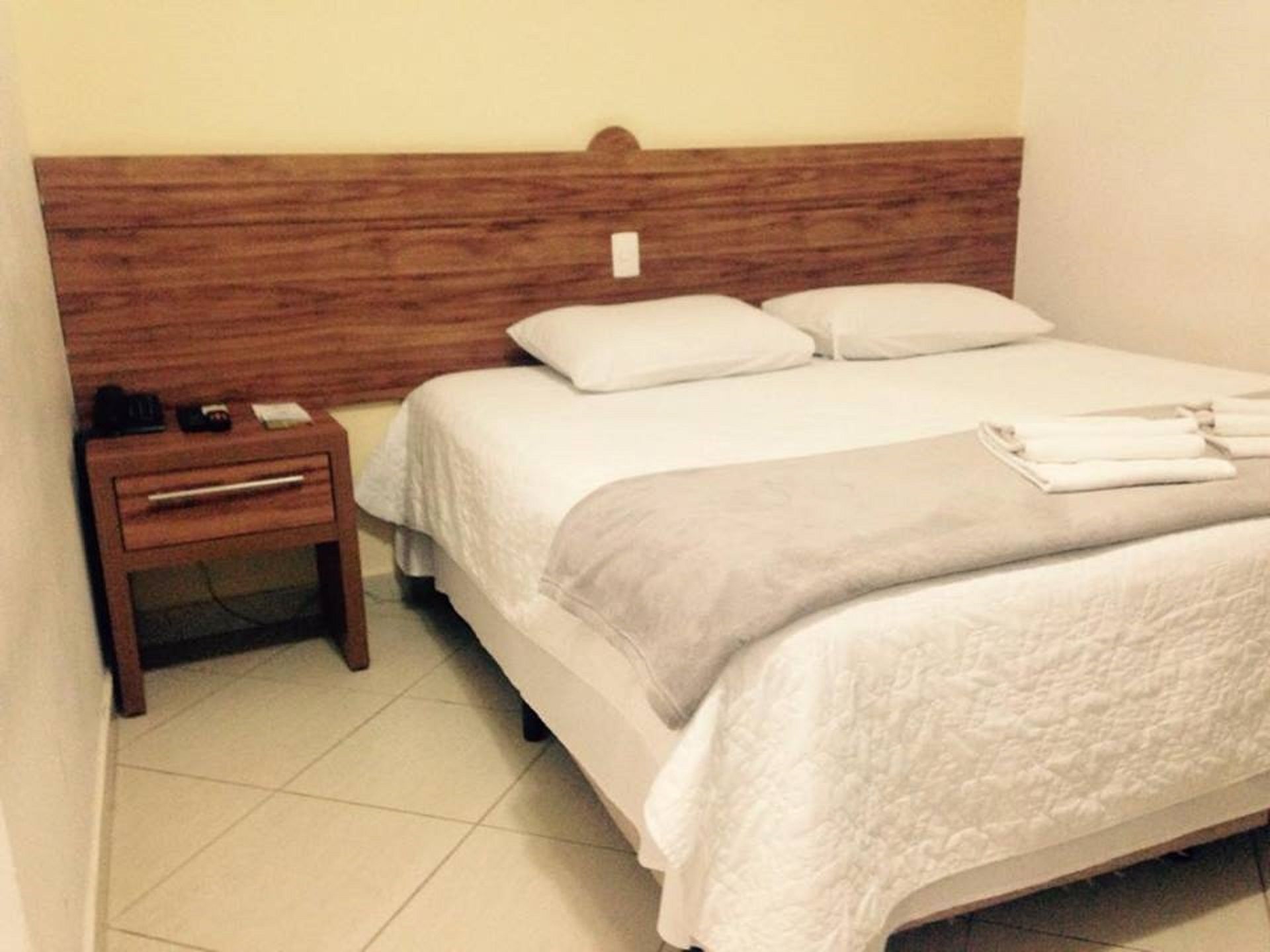 Cama bem arrumada com roupa de cama branca e um cobertor cinza, mesa de cabeceira de madeira com uma luminária, e uma parede lisa.